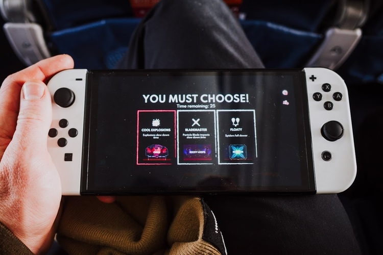 Mãos de um jogador segurando um console portátil Nintendo Switch OLED branco, exibindo uma tela de escolha de habilidades em um jogo com visual neon. No topo da tela, a frase "YOU MUST CHOOSE!".